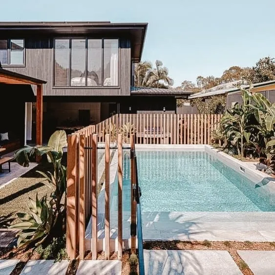 Piscine moderne avec une élégante clôture en bois, entourée de verdure et d'une maison contemporaine en arrière-plan.