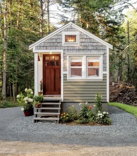 Mini-maison charmante avec une petite terrasse en bois, entourée de plantes fleuries et située dans une forêt tranquille.