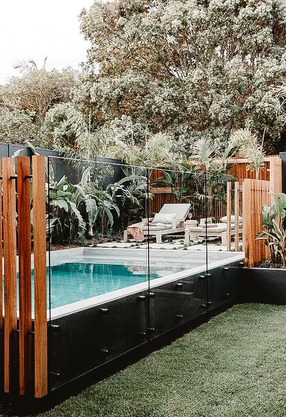 Piscine moderne avec une clôture en verre et poteaux en bois, entourée de végétation luxuriante et de chaises longues.