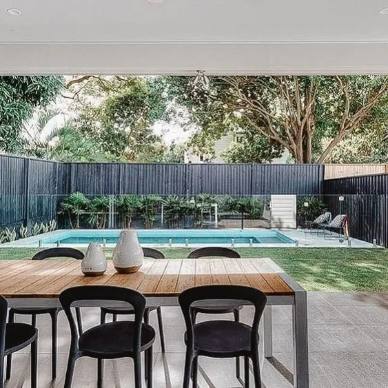 Modern pool area with a sleek wooden fence, surrounded by greenery and a contemporary home in the background.