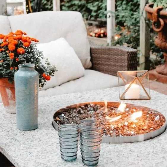 Cozy outdoor seating area with a fire bowl centerpiece on a table, surrounded by plants, candles, and stylish decor.