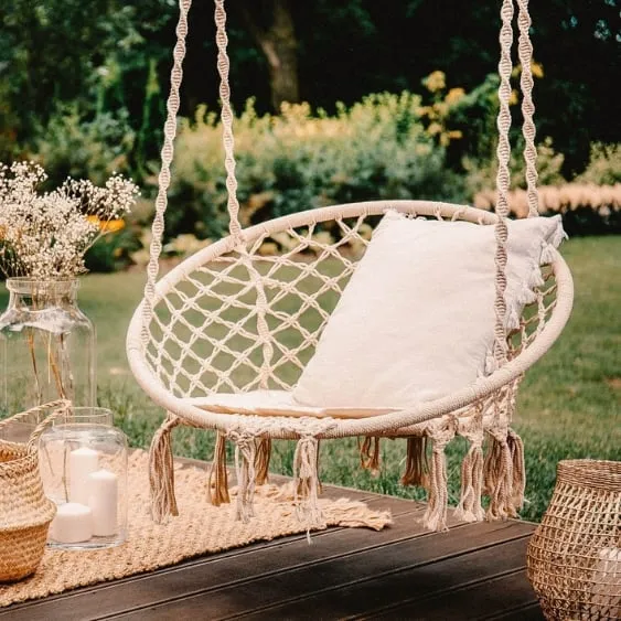 Macramé hanging chair with a cushion, surrounded by decorative items such as candles and vases, set on a lush outdoor deck.