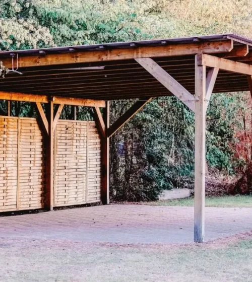 Wooden carport with a sturdy roof and privacy panels, set in a green, wooded backyard.