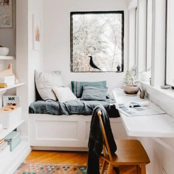 Coin lecture cosy et minimaliste avec un lit de jour et un bureau intégré, entouré d'étagères et baigné de lumière naturelle grâce aux grandes fenêtres, créant ainsi un refuge paisible.