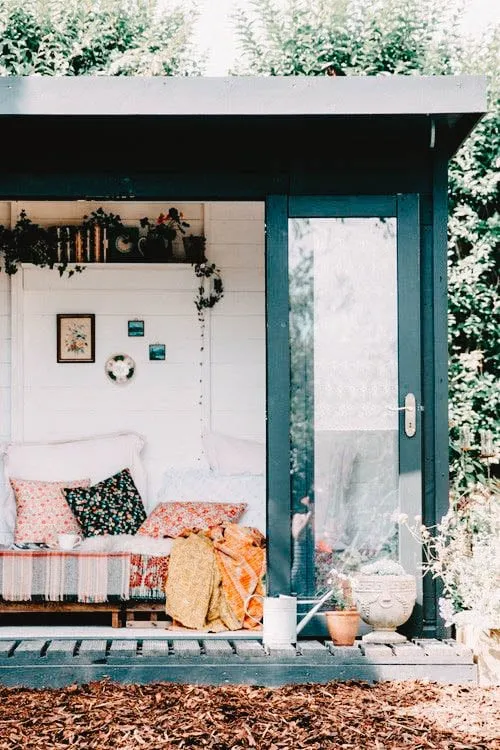 She Shed with a bohemian decor, featuring a cozy couch with colorful cushions, hanging plants, and a glass door, set in a lush garden.