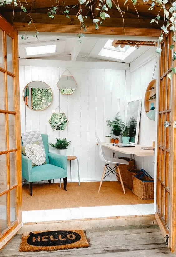 Cozy She Shed with a teal armchair, a small desk, and decorative mirrors on white walls, featuring natural light from skylights.