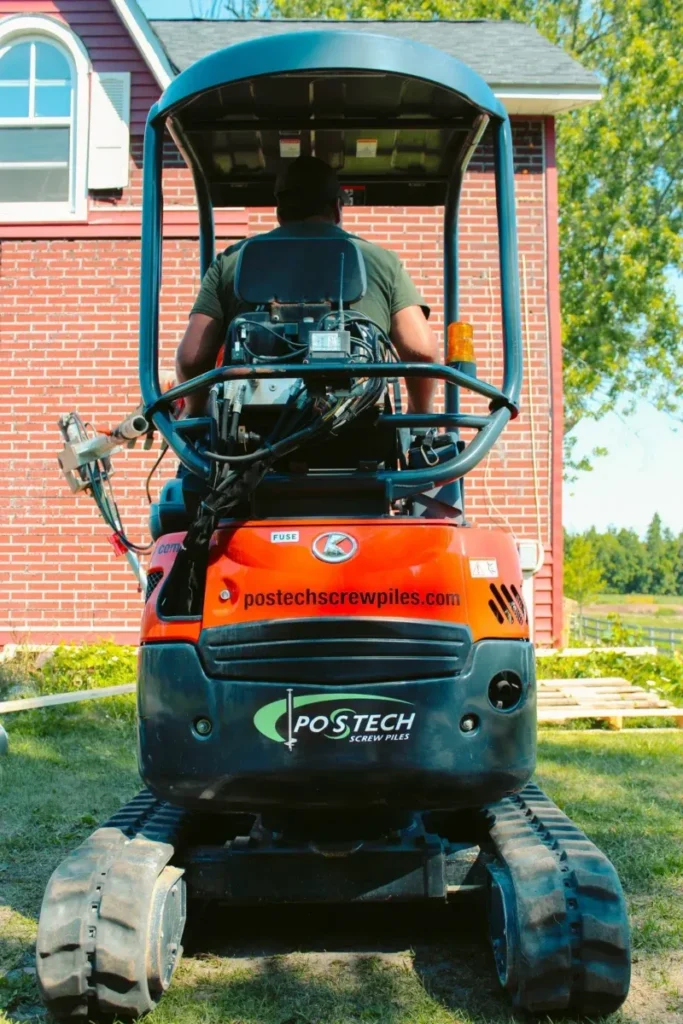 Back view of a worker operating a Postech screw pile machine near a brick house.