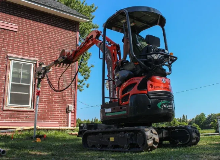 Back view of a worker operating a Postech screw pile machine near a brick house.