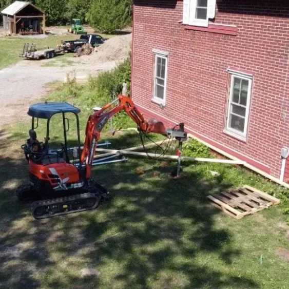 Excavator installing screw piles next to a brick house.