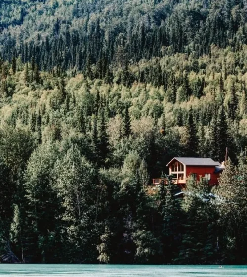 Secluded wooden cottage nestled in a dense green forest by a turquoise river, surrounded by mountainous landscape.