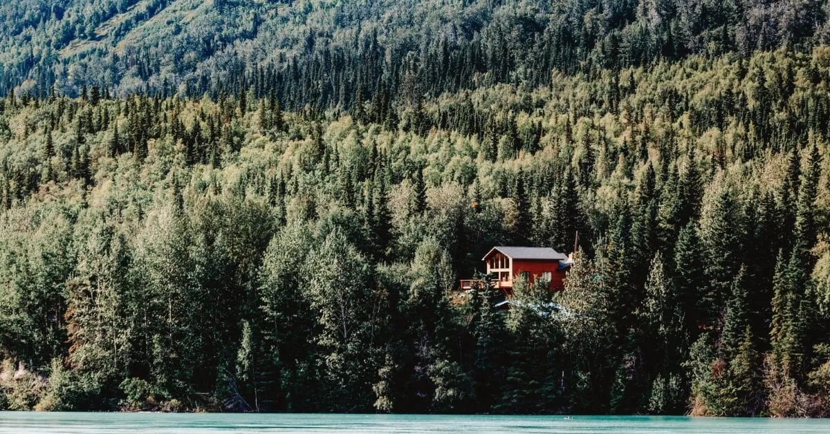 Secluded wooden cottage nestled in a dense green forest by a turquoise river, surrounded by mountainous landscape.