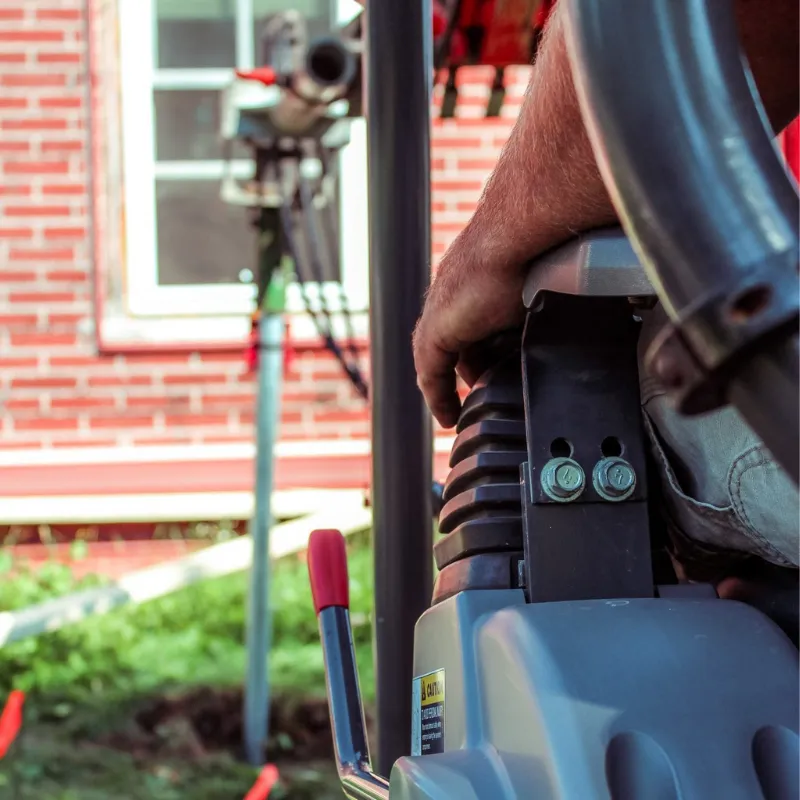 Close-up of a worker operating machinery for screw pile installation near a brick house.