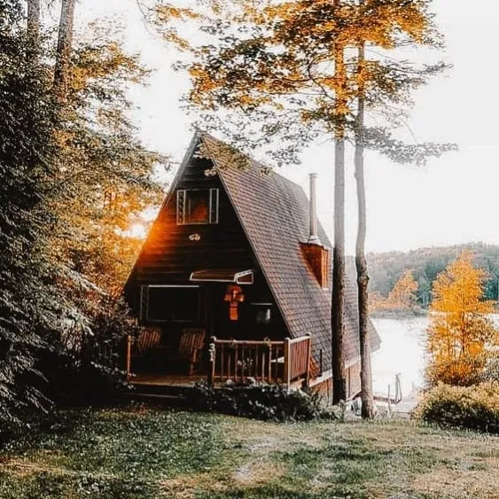 A-frame cabin nestled in a forest by a lake, surrounded by trees and bathed in golden sunset light.