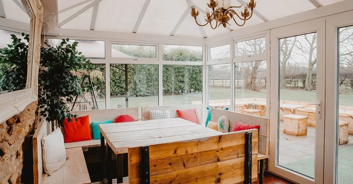 Cozy sunroom with large windows, wooden furniture, and colorful cushions, overlooking a garden.