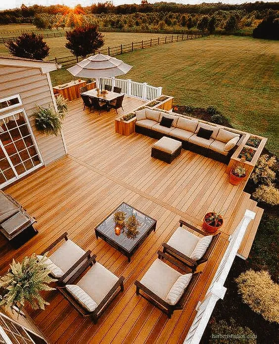 Large wooden deck with comfortable seating and lounging areas, overlooking a vast green field at sunset.