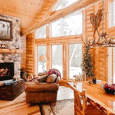 Rustic log cabin interior with a stone fireplace, wooden furniture, and large windows showcasing a snowy outdoor landscape.