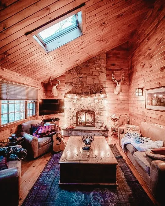 Warm cabin interior with a stone fireplace, wooden walls, cozy seating, and natural light from a skylight, creating a peaceful and inviting atmosphere.