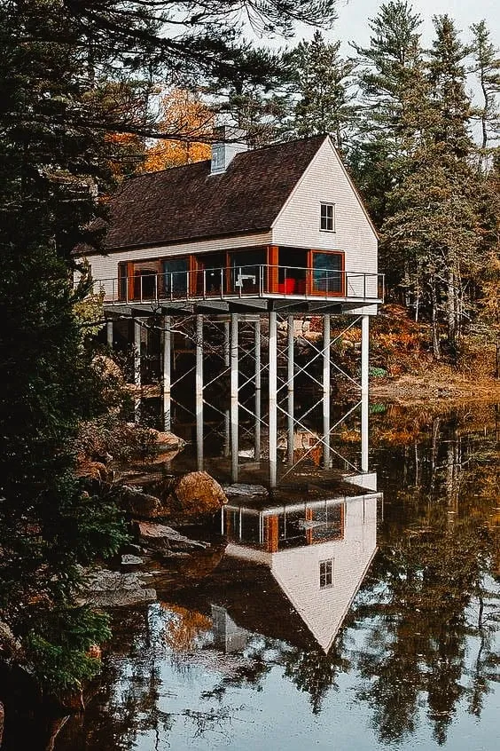 House elevated on stilts over a calm lake, surrounded by dense forest, with a perfect reflection on the water.