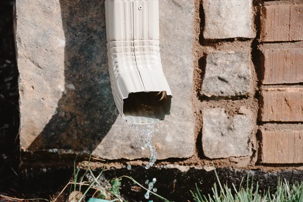 Water flowing out of a gutter downspout near the foundation of a home, set against a stone and brick exterior wall.