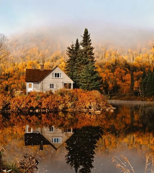 A cozy wooden house surrounded by autumn trees, reflecting on a calm lake, with mist-covered hills in the background.