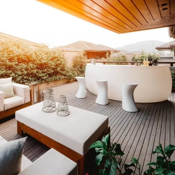 Modern outdoor deck with comfortable seating, a sleek bar, and lush greenery, bathed in warm sunlight.