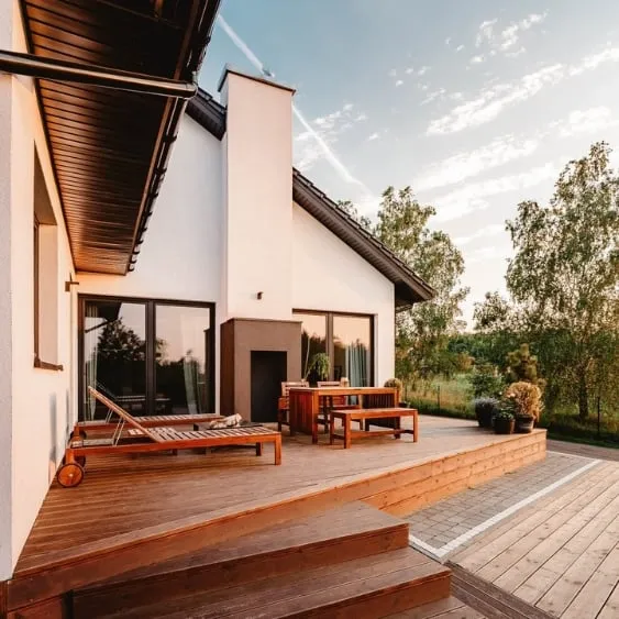 A modern home with a spacious wooden deck featuring outdoor furniture, surrounded by lush greenery and a clear sky, perfect for entertaining.