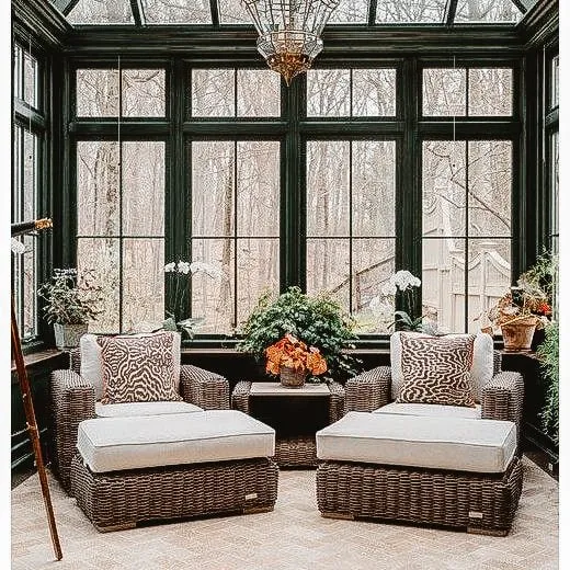 Cozy sunroom with large windows overlooking a forest, featuring two comfortable wicker lounge chairs with plush cushions and a central table adorned with potted plants.