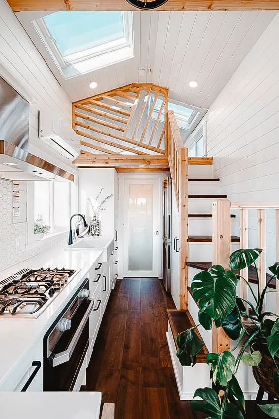 Bright and modern tiny home interior with wooden accents, featuring a compact kitchen, skylights, and a loft accessible by a wooden staircase.