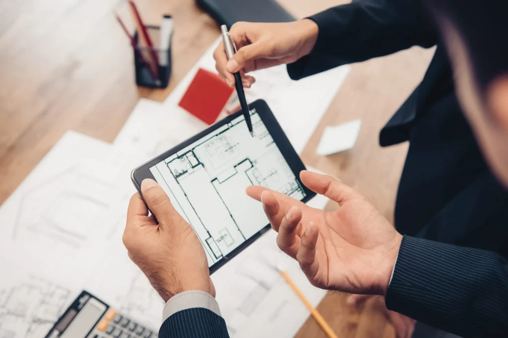 Two professionals discussing architectural plans on a tablet, surrounded by blueprints and design materials, illustrating effective planning in construction.