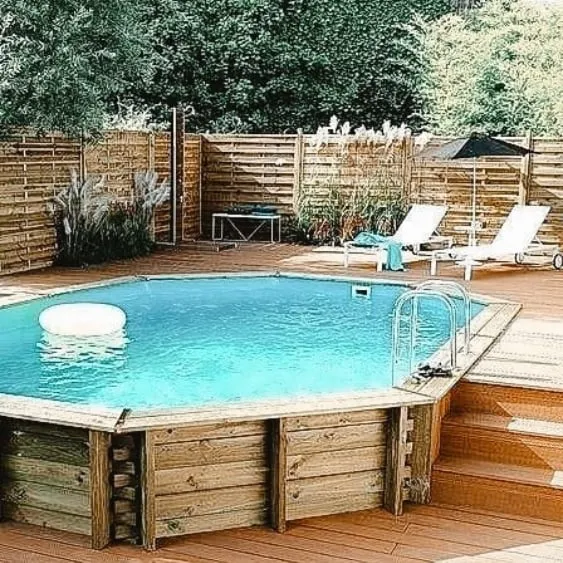 Terrasse de piscine en bois avec une piscine hors-sol octogonale et des chaises longues, entourée d'une clôture en bois dans un cadre verdoyant.