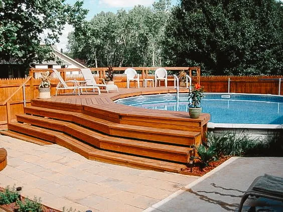 Wooden deck surrounding an above-ground pool with multi-level steps and lounging chairs, set in a private backyard with greenery.