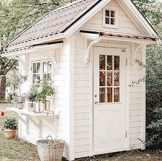 Small white shed with a glass-paneled door and gable roof, surrounded by greenery and decorated with potted plants.