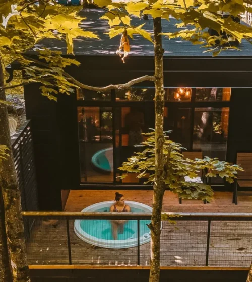 Aerial view of a modern cabin in the woods with a deck and hot tub, built on screw piles.