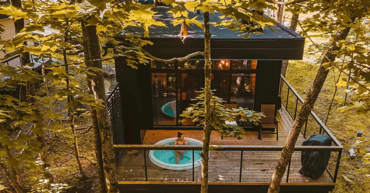 Aerial view of a modern cabin in the woods with a deck and hot tub, built on screw piles.