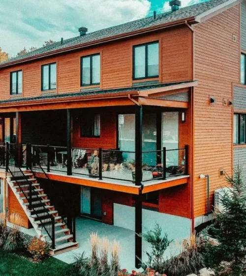 Two-story modern residential buildings with orange siding and glass-railed balconies, supported by black screw pile foundations.