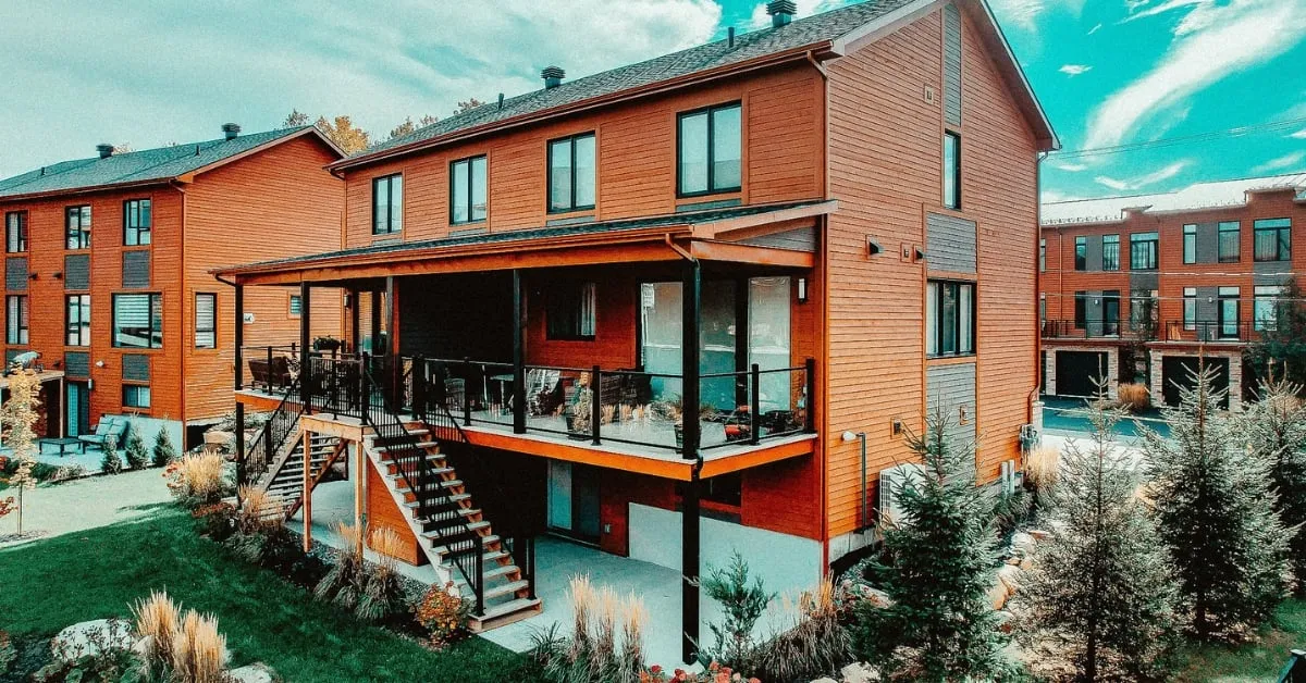 Two-story modern residential buildings with orange siding and glass-railed balconies, supported by black screw pile foundations.