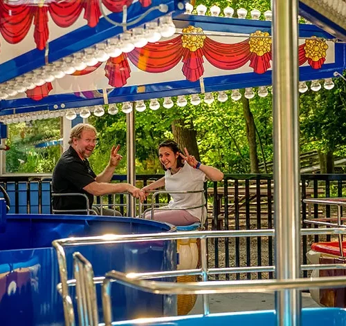 Two people enjoying a colorful amusement ride at Santa's Village, surrounded by lush greenery in a family-friendly outdoor setting.