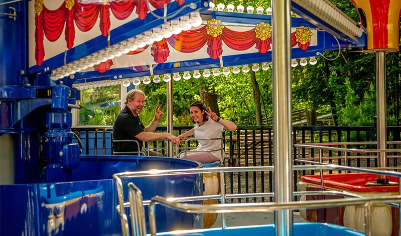 Two people enjoying a colorful amusement ride at Santa's Village, surrounded by lush greenery in a family-friendly outdoor setting.