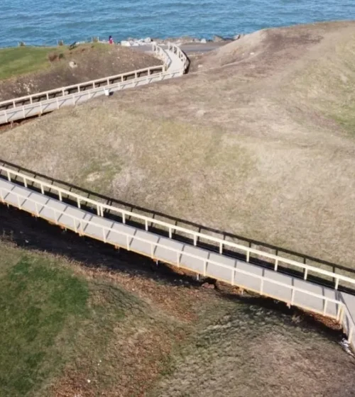 Aerial view of a wooden boardwalk supported by screw piles, winding along a historical trail by the water's edge.