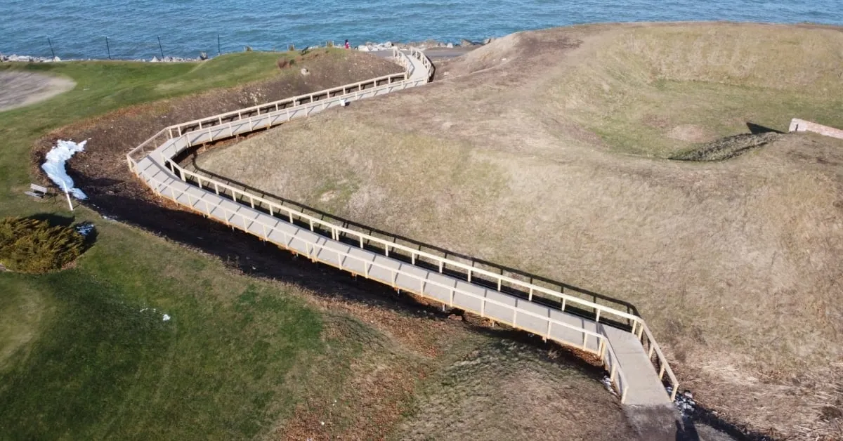 Aerial view of a wooden boardwalk supported by screw piles, winding along a historical trail by the water's edge.