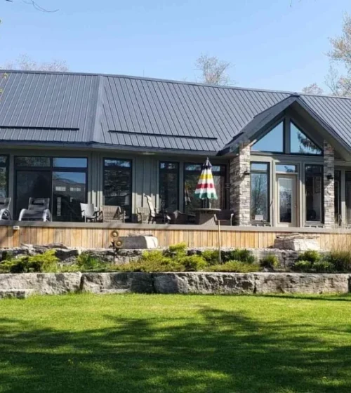 Modern single-story house with a sleek metal roof, large glass windows, and a spacious stone-lined patio, surrounded by lush greenery and trees.