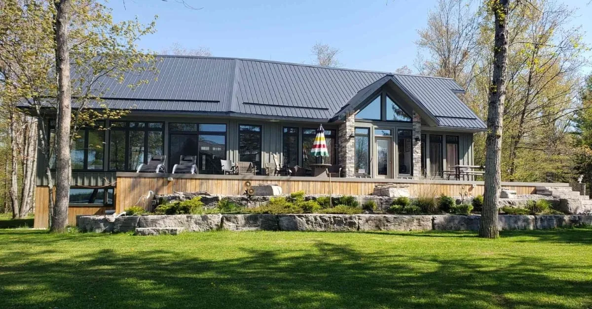 Modern single-story house with a sleek metal roof, large glass windows, and a spacious stone-lined patio, surrounded by lush greenery and trees.