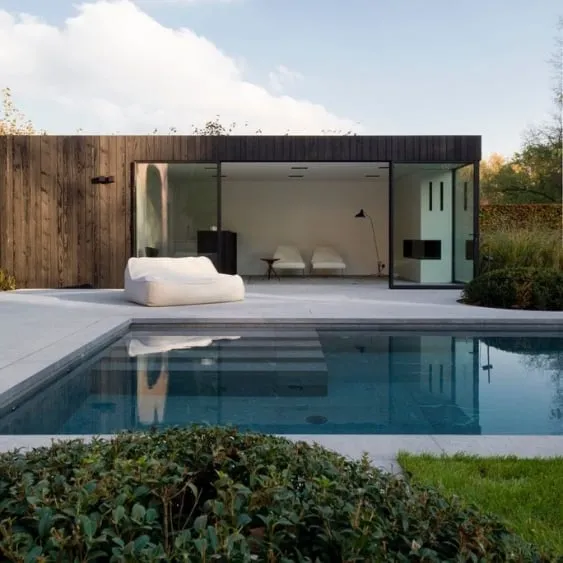 A modern pool house with large glass windows and a sleek design, surrounded by greenery and a serene pool, perfect for relaxation.