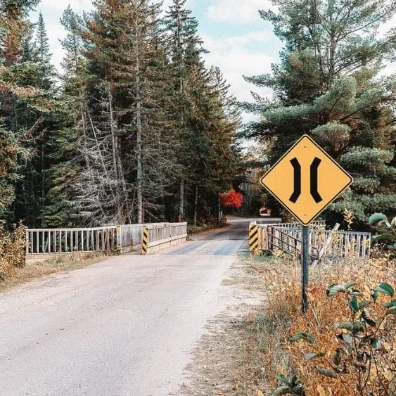 A road sign on a quiet rural road with a bridge, supported by screw piles, amidst a scenic forest setting.