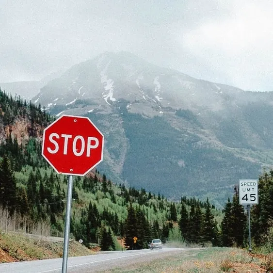 Stop sign along a scenic road with a mountainous backdrop and trees lining the roadside.