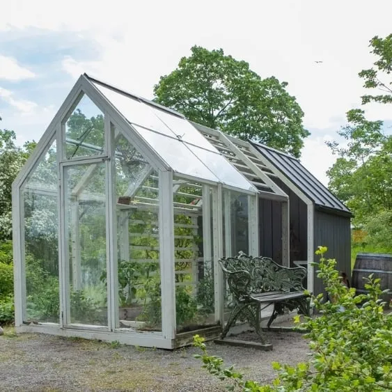 Small greenhouse with glass panels and a metal bench outside, surrounded by greenery in a garden setting.