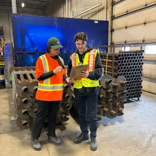 Two Postech Screw Piles employees in safety gear discussing work in an industrial warehouse setting with metal pipes in the background.