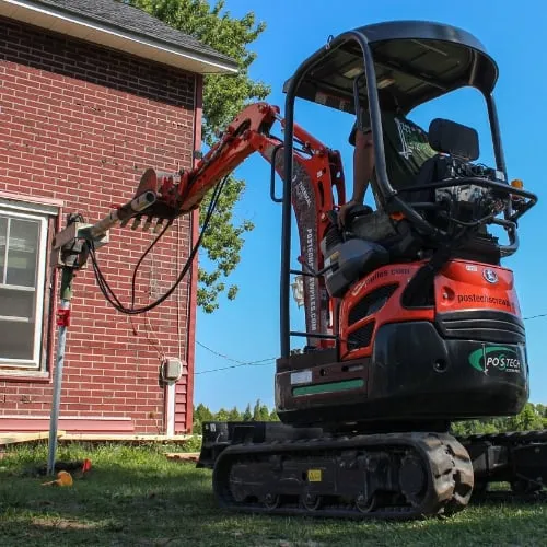 Orange mini-excavator installing a screw pile near a red brick house.