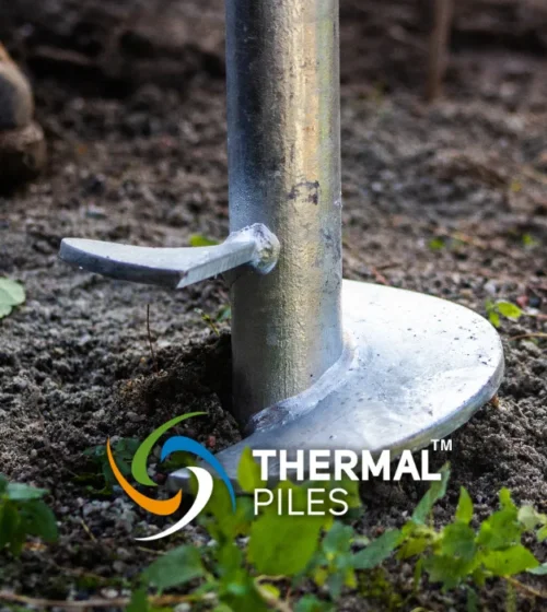Close-up of a metal screw pile embedded in the soil with the "Thermal Piles" logo and a worker's boot partially visible in the background.