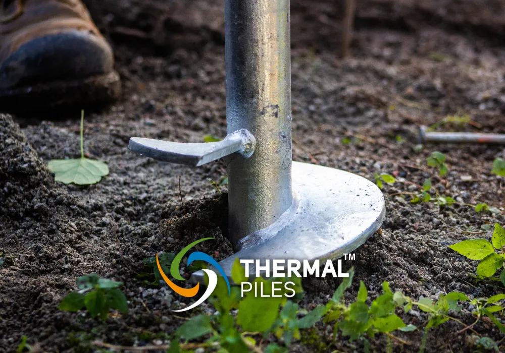 Close-up of a metal screw pile embedded in the soil with the "Thermal Piles" logo and a worker's boot partially visible in the background.